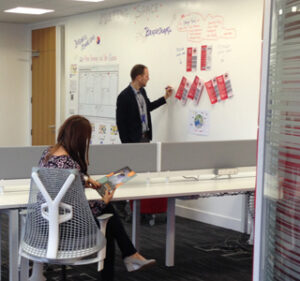 Man presenting at whiteboard, woman seated and reading in a modern office space.