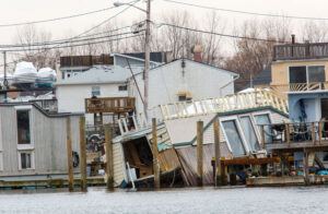 Flood-damaged houses tilted and partially submerged in water near a shoreline.