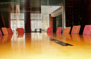 Empty conference room with a long wooden table and red chairs, surrounded by large windows with blinds.