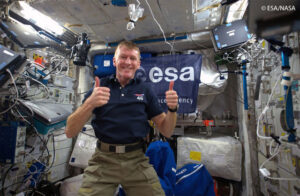 Astronaut giving thumbs up inside the International Space Station with ESA banner in the background.