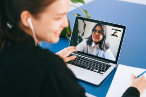 A woman smiling during a video call on a laptop with another woman, while taking notes on a piece of paper.