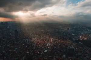 Aerial view of a vast city at sunset, with sunbeams breaking through dramatic clouds over densely packed buildings.