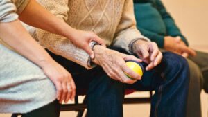 A person gently holds the arm of an elderly person squeezing a stress ball, while another person sits beside them.