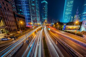 Cityscape at night with light trails on busy roads, surrounded by tall illuminated buildings.