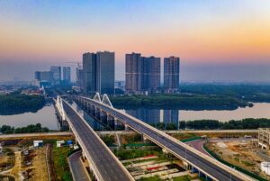 Aerial view of a bridge over a river with tall buildings and lush greenery on a clear day.