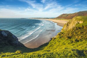 Coastal landscape with green cliffs, a sandy beach, and waves under a blue sky.