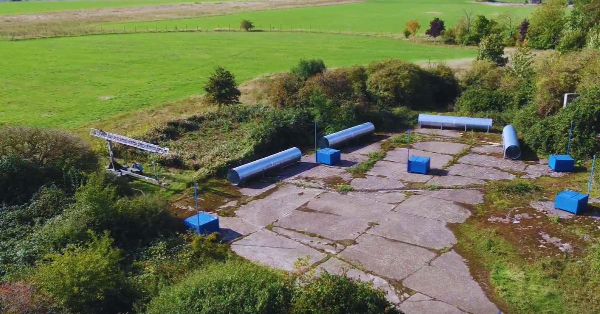 Aerial view of an abandoned concrete site with blue boxes and large cylindrical metal pipes, surrounded by grass.