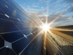 Close-up of solar panels with the sun shining brightly in a blue sky with some clouds.