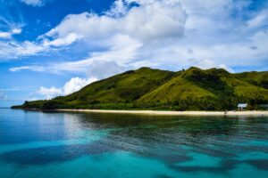 A tranquil island with lush green hills, a sandy beach, and clear blue waters under a partly cloudy sky.