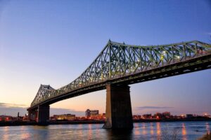 Illuminated green steel bridge spans over a calm river at dusk, with city lights twinkling in the background.