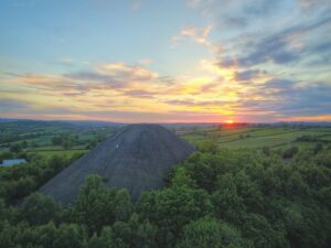 Sun setting over a green landscape with a large mound, surrounded by lush trees and fields.