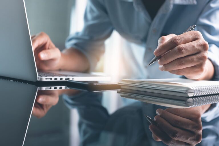 Person writing in a notebook while using a laptop on a glass desk.
