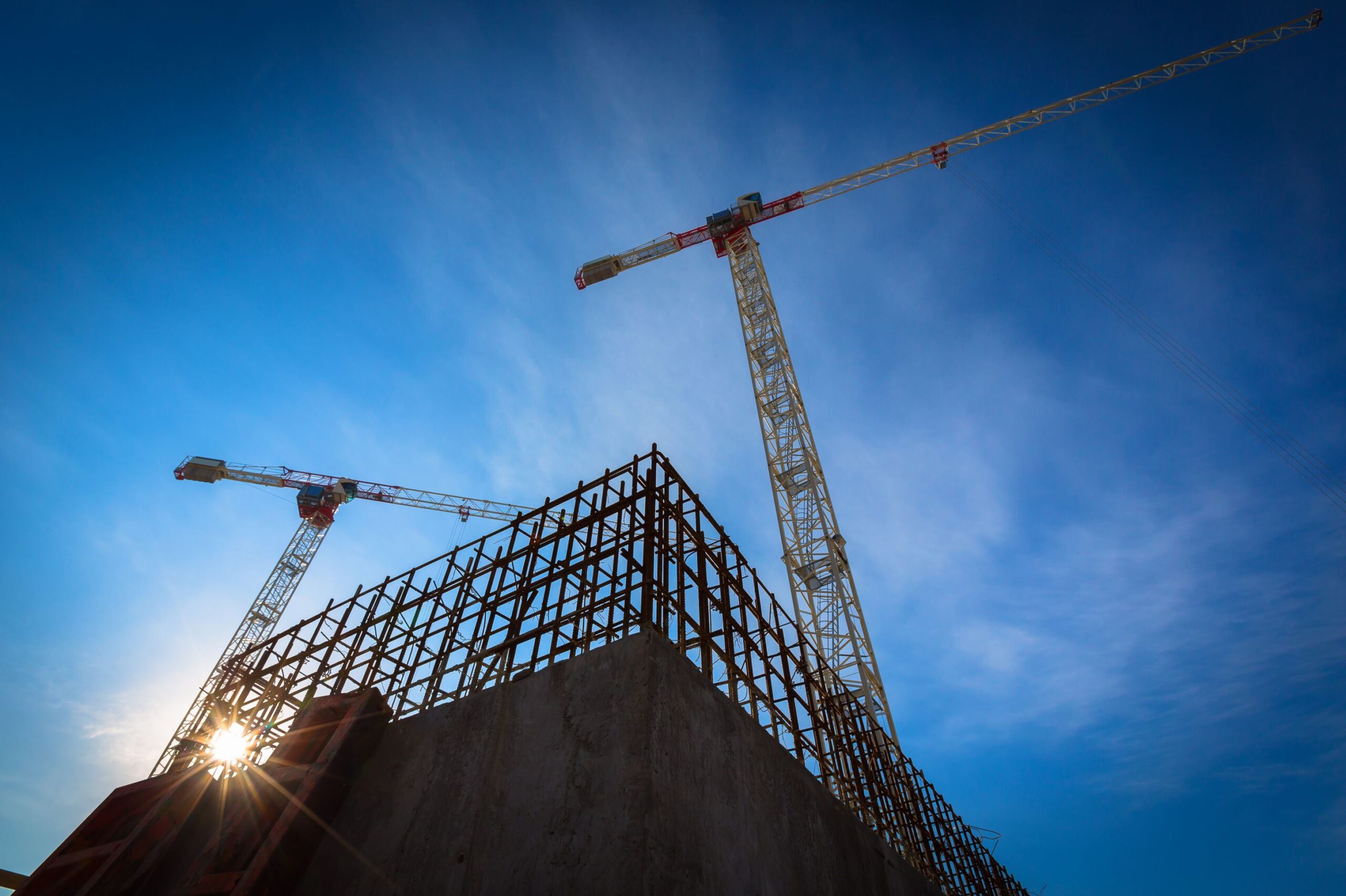 Construction site with two tall cranes and steel framework silhouetted against a clear blue sky.