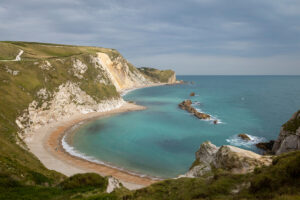 A coastal landscape with cliffs, a sandy beach, and turquoise water under a cloudy sky.