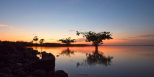 Sunset over a calm lake with silhouetted trees and a rocky shoreline, reflecting warm orange and blue hues.