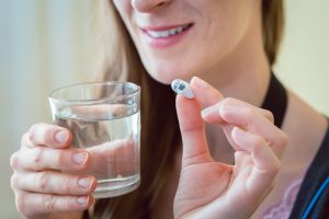 A woman smiles, holding a capsule and a glass of water.