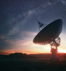 Silhouetted satellite dish against a starry sky at dusk, with distant hills on the horizon.