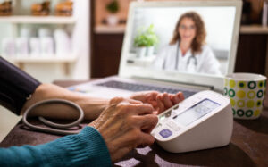 Person using a digital blood pressure monitor during an online consultation with a doctor on a laptop screen.