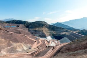 Open-pit mine with large terraced slopes, excavators, and mountainous backdrop under a clear blue sky.