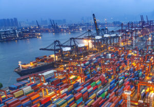 Colorful shipping containers stacked at a busy port, with cranes and a ship, city skyline visible in the distance.