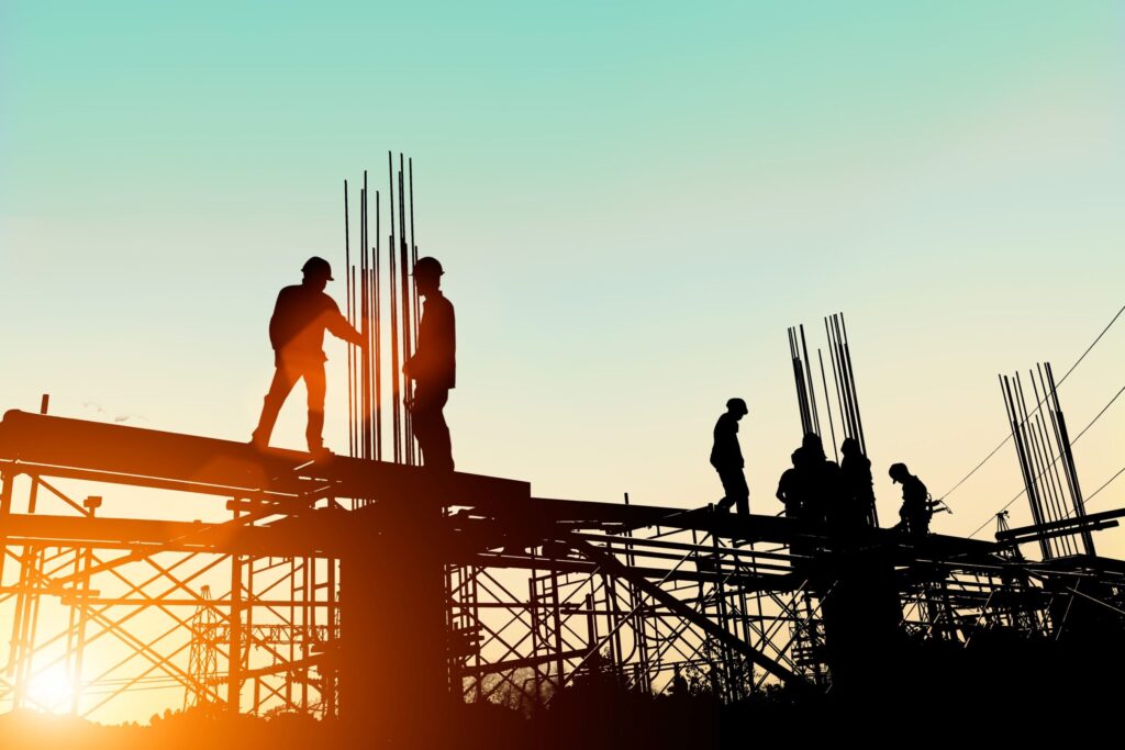 Silhouettes of construction workers on scaffolding at sunset, with a colorful sky in the background.