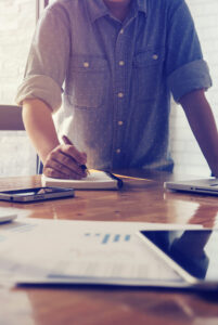 Person in a denim shirt writing in a notebook at a table with laptops, smartphones, and documents.