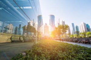 Modern city skyline with glass buildings, escalators, and sunlit skyscrapers reflecting in a window. Greenery in foreground.