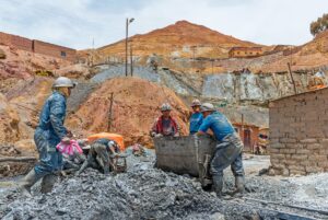 Miners in helmets working at a rocky site with carts, dirt piles, and distant buildings under a cloudy sky.