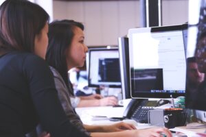 Two women working at a computer in an office setting, focusing on the screen.