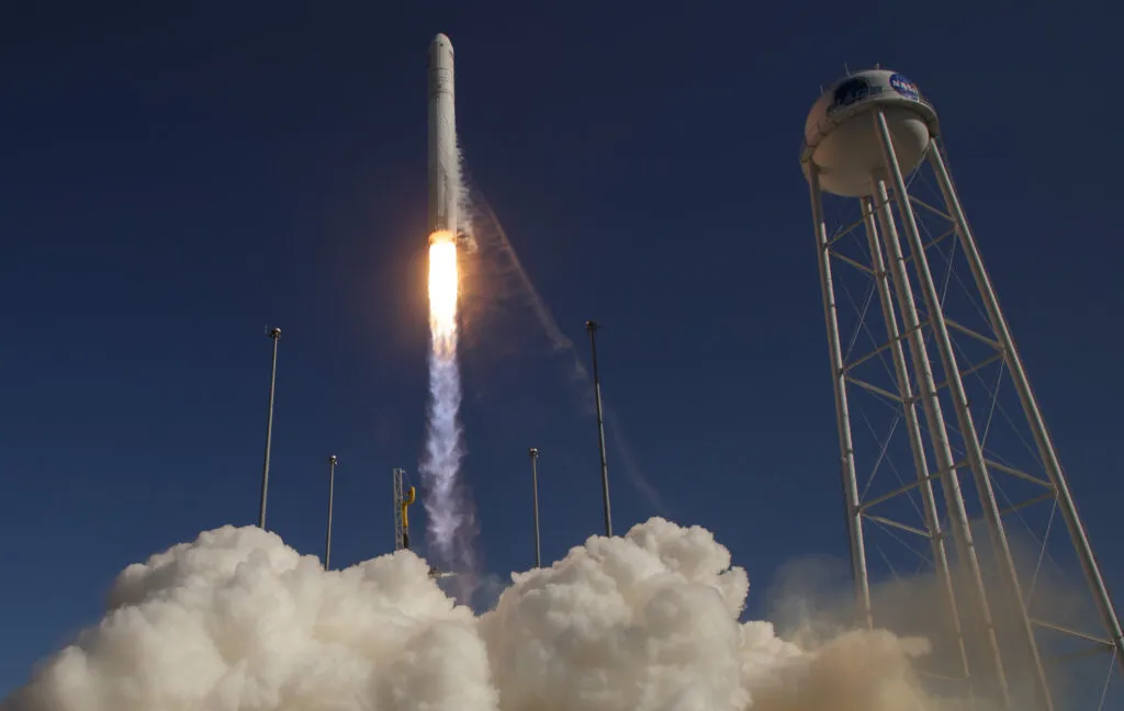A rocket launches into a clear blue sky, with clouds of smoke at its base and a tall water tower nearby.