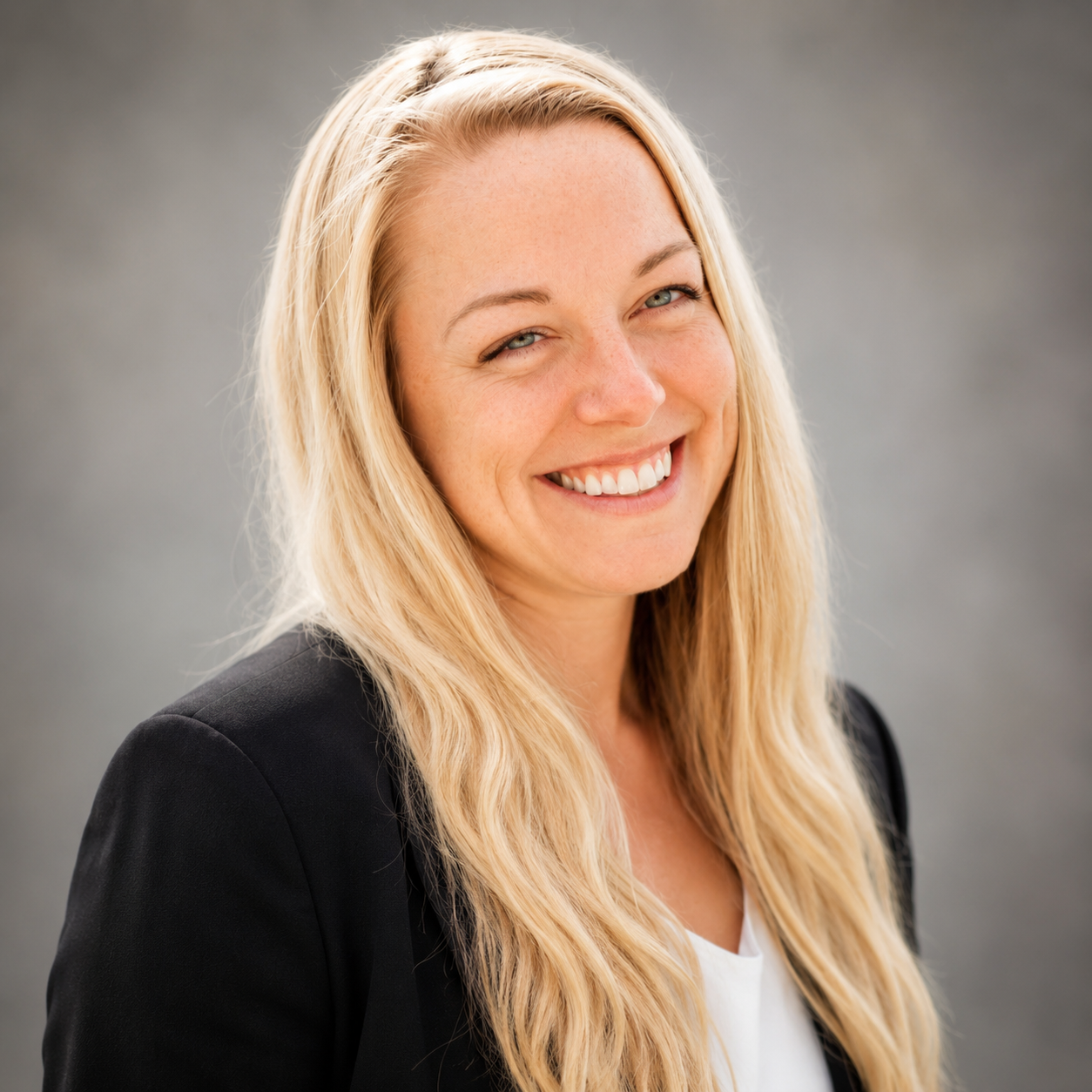 Woman with long blonde hair in a black blazer smiling in front of a gray blurred background.