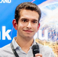 Man with dark hair holding a microphone, standing in front of a blue background with white text.