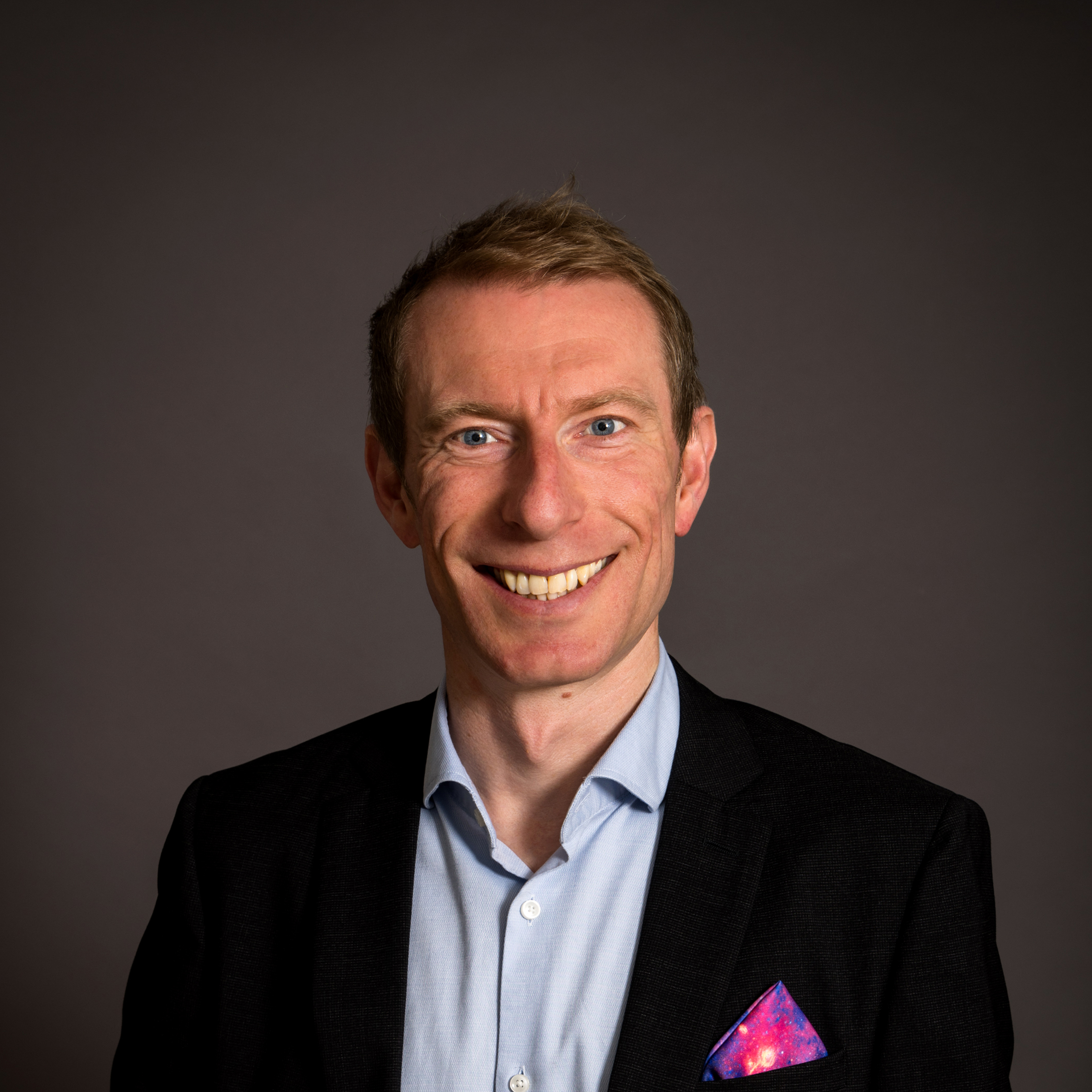 Smiling man in a suit jacket with a colorful pocket square, standing against a plain dark background.