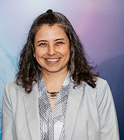 A woman with wavy dark hair, wearing a light gray blazer and patterned shirt, smiling at the camera.