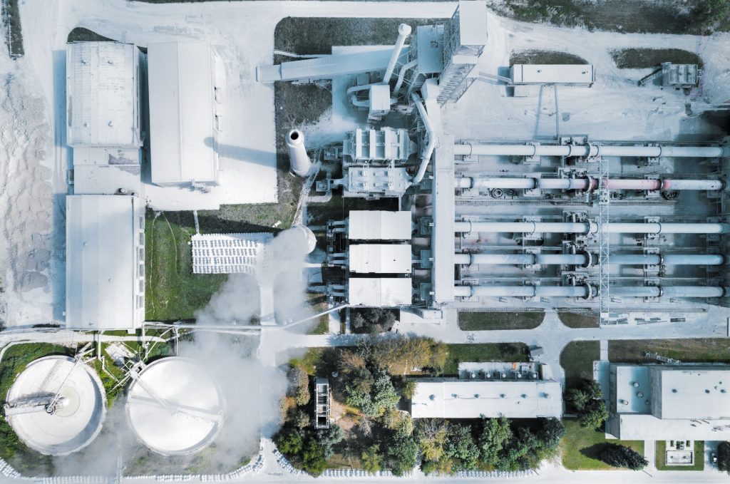 Aerial view of an industrial facility with numerous buildings, smokestacks, and storage tanks surrounded by trees.