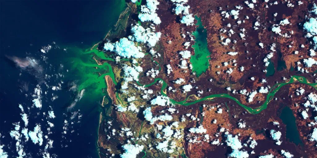 Satellite view of a winding green river, land, and clouds next to a dark blue ocean.
