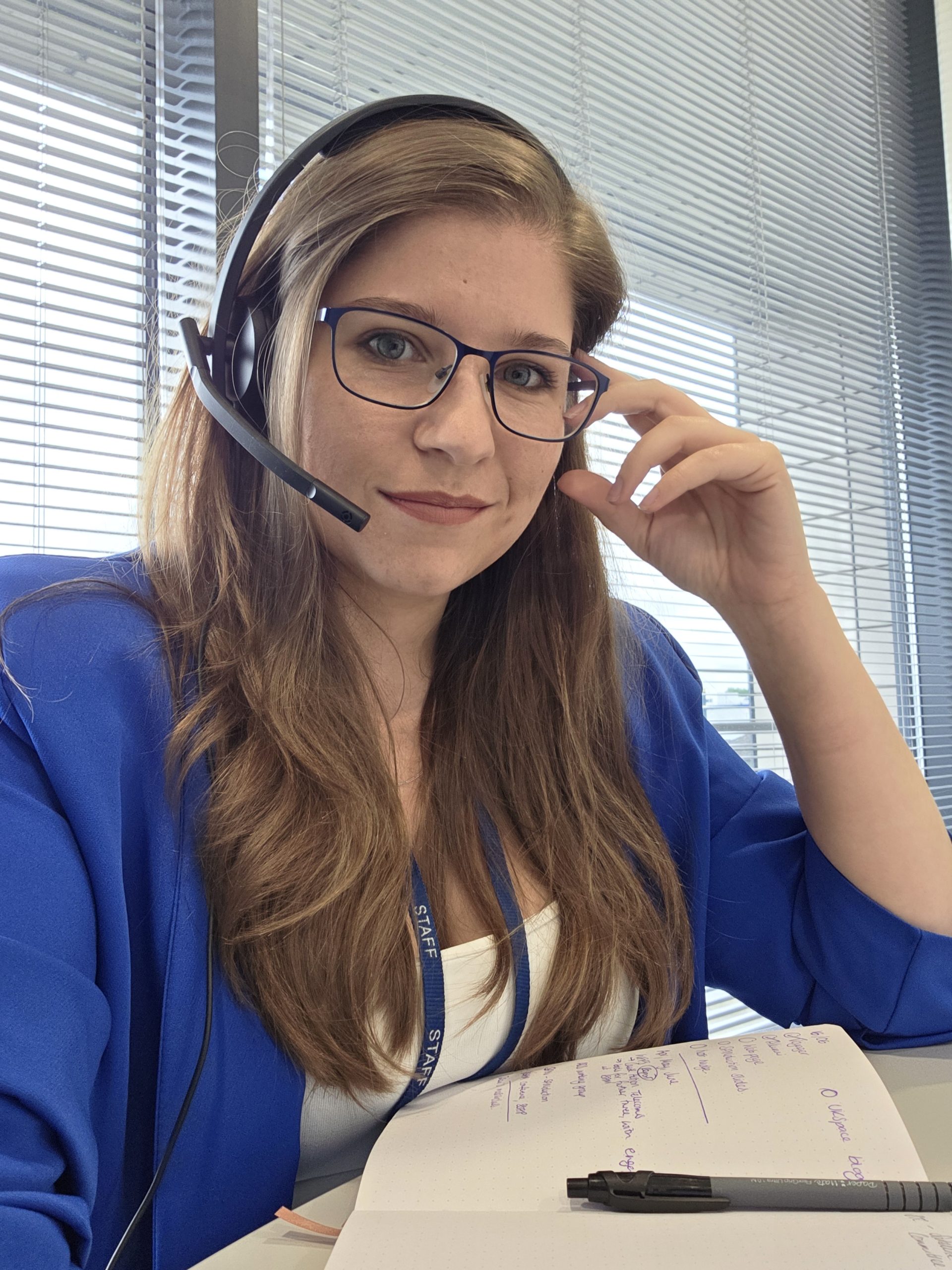 A woman with glasses and a headset sits at a desk with a notebook, smiling at the camera.