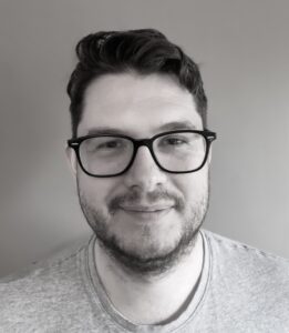 Man with glasses and a beard smiles slightly, wearing a gray t-shirt. Gray background.