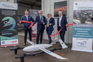 Four people celebrate a grand opening next to a drone and banners outside a building.