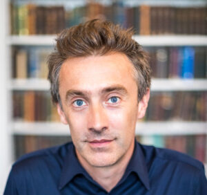 Person with short brown hair and blue eyes wearing a navy shirt, standing in front of a blurred bookshelf background.