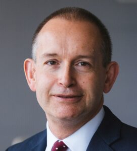 A man in a suit and tie smiles against a plain background.
