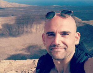 A person smiles at the camera with a desert landscape and crater in the background. Sunglasses rest on their head.