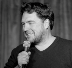 A person smiling and holding a microphone, speaking on stage in a black-and-white photo.