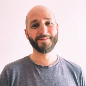 A man with a beard smiles softly at the camera, wearing a gray shirt against a plain light background.