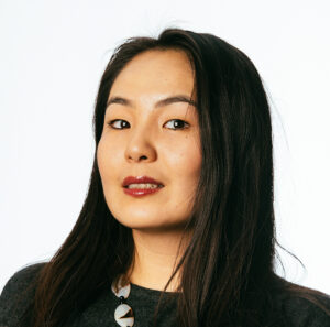 Person with long dark hair and a necklace, looking at the camera against a plain white background.
