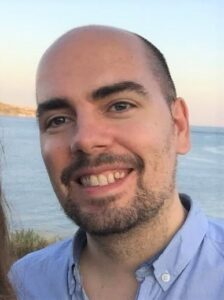 A smiling man with a beard stands near the sea, wearing a light blue shirt. The background shows a calm ocean and sky.