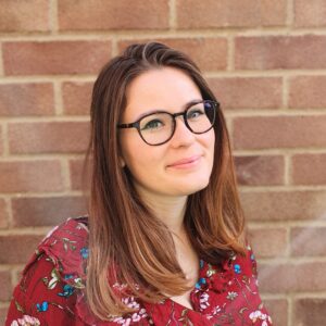 A woman with glasses and long hair smiles, wearing a floral top, standing in front of a brick wall.