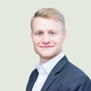 A person with short blonde hair and a suit smiles at the camera against a light gray background.
