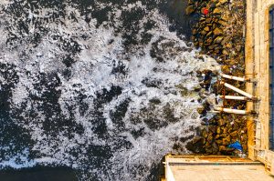 Aerial view of foamy water discharge from pipes flowing into a body of water, surrounded by rocks.