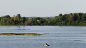 Small boat with two people on a wide river, surrounded by trees and distant greenery under a clear sky.